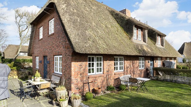 Une maison en briques avec un toit de chaume, une terrasse dotée de mobilier d’extérieur et un jardin bien entretenu.
