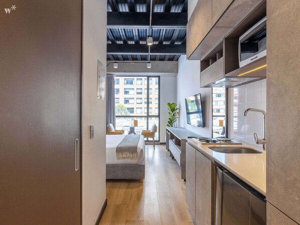 Een moderne hotelkamer met een kitchenette, een groot raam met uitzicht op de stad, een zithoek met een bank en een hoog plafond met zichtbare balken.