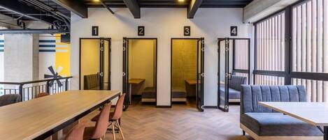 A modern interior with a long wooden table, multiple chairs, and a sofa. There are four numbered doors leading to private rooms.