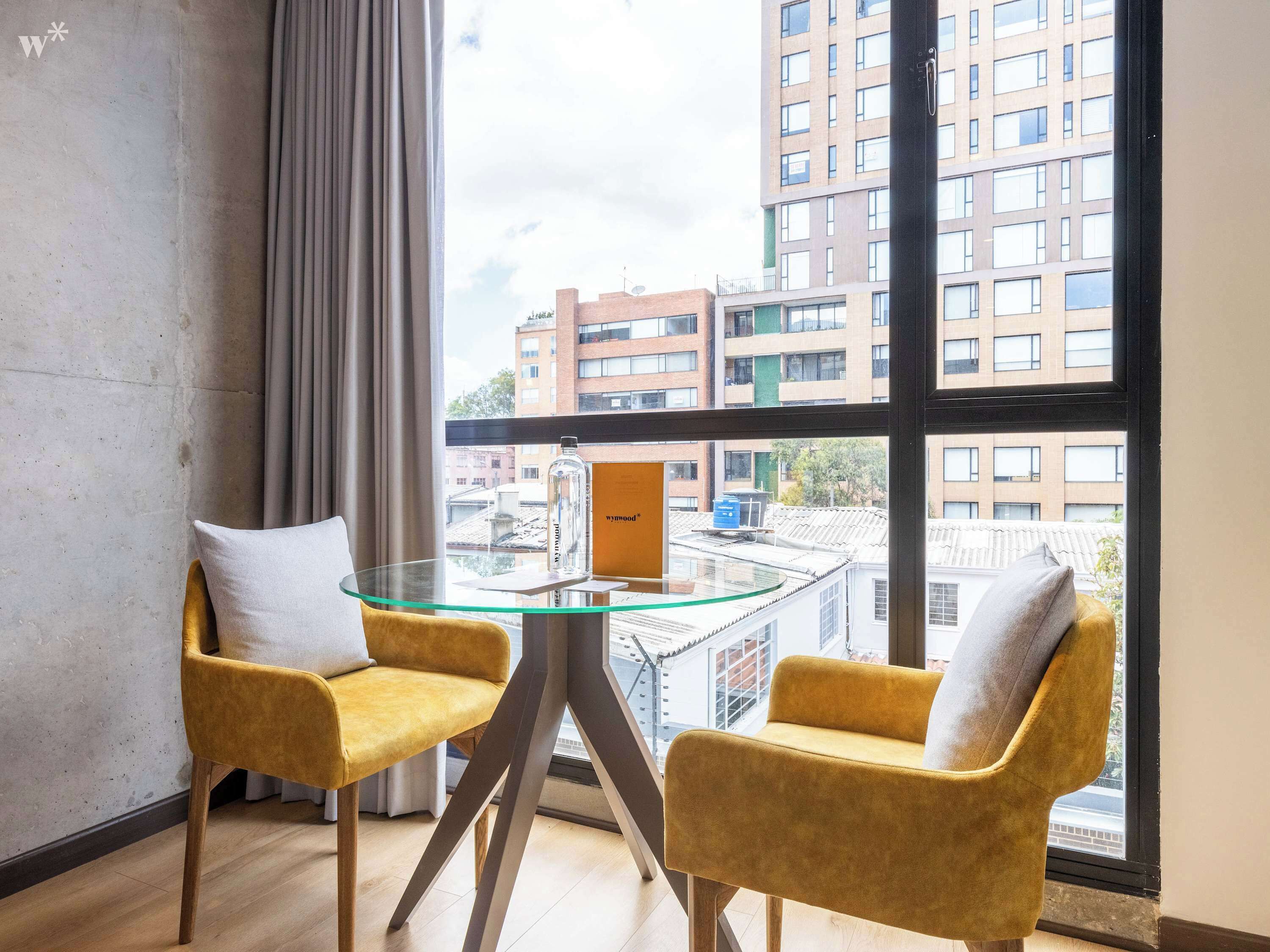 A modern room with two yellow chairs, a glass table, and a large window offering a view of buildings.