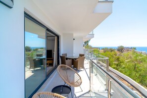 Ein Balkon mit Glastüren, Korbstühlen, einem Tisch und Blick auf das Meer.