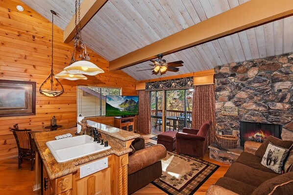 A log cabin interior with a stone fireplace, wooden furniture, and a kitchen area.