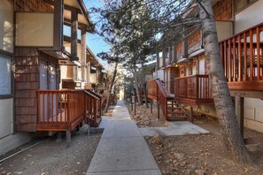 A narrow pathway flanked by wooden buildings with balconies, a tree-lined path, and a clear sky.