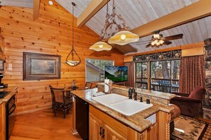 A wooden cabin interior with a kitchen, dining area, and a large window overlooking a deck.