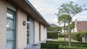 Un bâtiment moderne avec une façade en briques, de grandes fenêtres et un jardin bien entretenu agrémenté de haies et d’un arbre.