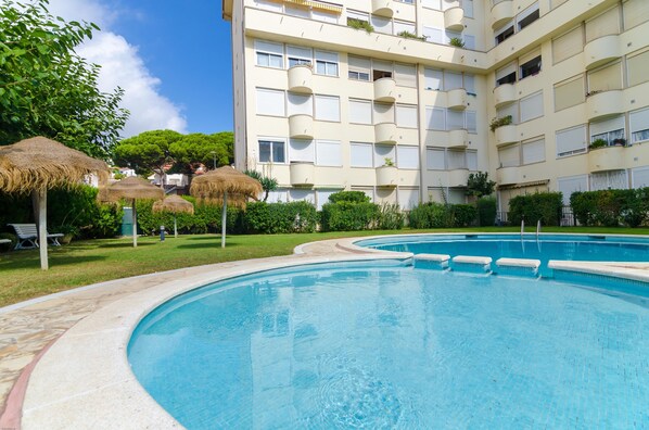 A swimming pool with a curved edge, surrounded by a grassy area with thatched umbrellas. - Apartamento Majestic (Lloret de Mar)