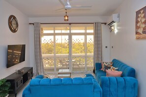 A living room with a blue sofa, a flat-screen TV, a ceiling fan, and a large window with a view of greenery.