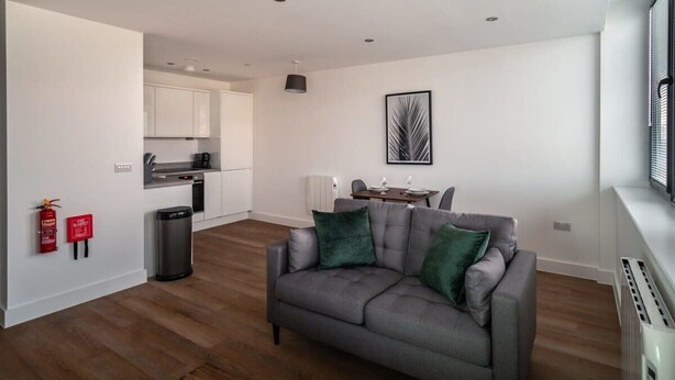 A modern living room with a grey sofa, a dining table with chairs, a kitchen area, and a fire extinguisher on the wall. - Stay Zeni, Alexandra Rise Manchester Modern 1 Bed (Manchester)