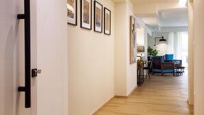 A hallway with framed pictures on the wall, a dining area with chairs and a table, and a living room with a sofa and a lamp.