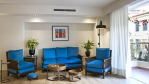 A living room with a blue sofa set, two side tables with potted plants, a floor lamp, and a large window with curtains.