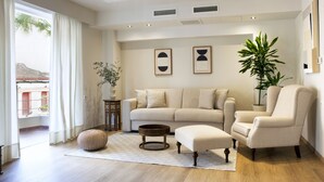 A living room with a beige sofa, a round coffee table, and a beige armchair. There is a large potted plant, a small side table with a vase, and artwork on the wall.