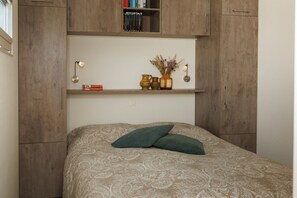 A bedroom with a bed, a shelf with books and decorative items, and a wall-mounted light.