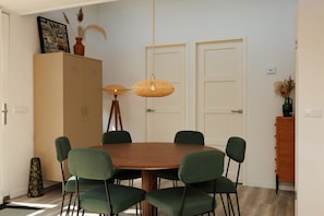 A dining area with a round wooden table, six green chairs, two white doors, and a cabinet with a picture frame.