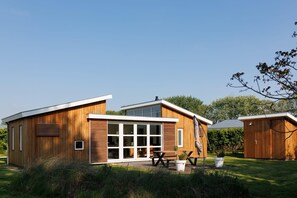 A modern wooden house with a large glass door entrance, a patio with a table and chairs, and a small shed in the background.