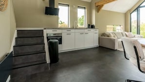 A modern kitchen with a built-in oven, a sink, and a window. There is a staircase leading to another level, and a seating area with a sofa and chairs.
