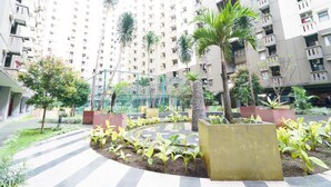 Un immeuble résidentiel doté de nombreux balcons, une cour aménagée avec des palmiers et un chemin bordé d’arbres disposés en damier.