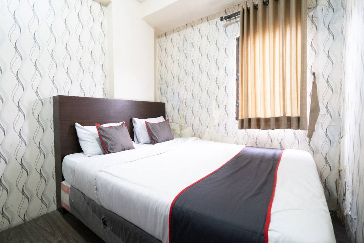 A hotel room with a large bed, a wooden headboard, patterned wallpaper, and a window with curtains.