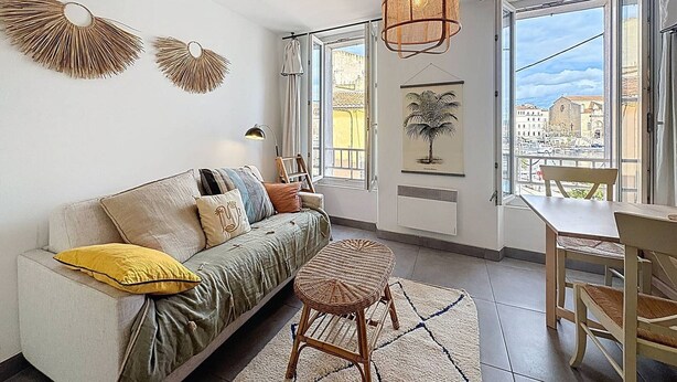 A modern living room with a sofa, a dining table, and chairs. There are large windows with a view of buildings and a cityscape. - Bright T2 Apartment at the Old Port, La Ciotat (La Ciotat)