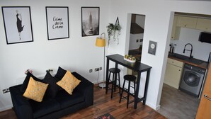 Ett modernt vardagsrum med en svart soffa, gula kuddar, ett litet bord med en krukväxt och en köksdel med en tvättmaskin.