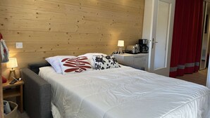 A hotel room with a wooden headboard, a bed with white bedding and decorative pillows, a bedside table with a lamp and a coffee maker, and a red curtain.