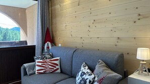 A modern living room with a grey sofa, patterned cushions, a wooden wall, and a window with a view of trees.