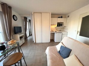 Ein kompakter Wohnbereich mit Küchenzeile, einer Couch mit Kissen, einem Esstisch mit Stühlen, einem Fernseher und einer Wanduhr.