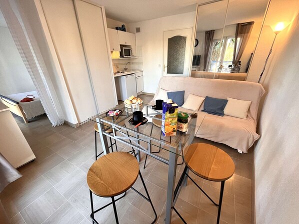 A modern living room with a glass dining table, two wooden stools, a sofa, and a kitchen in the background.