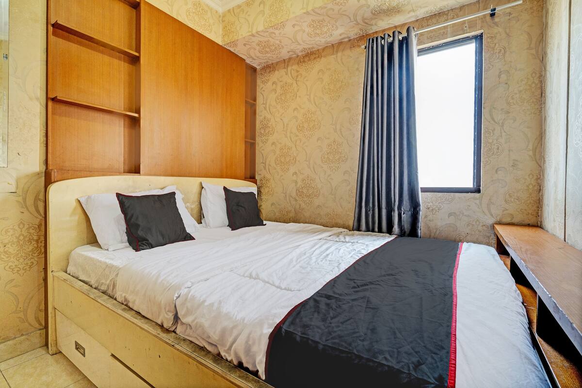 A bedroom with a bed, wooden headboard, built-in shelves, a window with curtains, and a wooden bench.