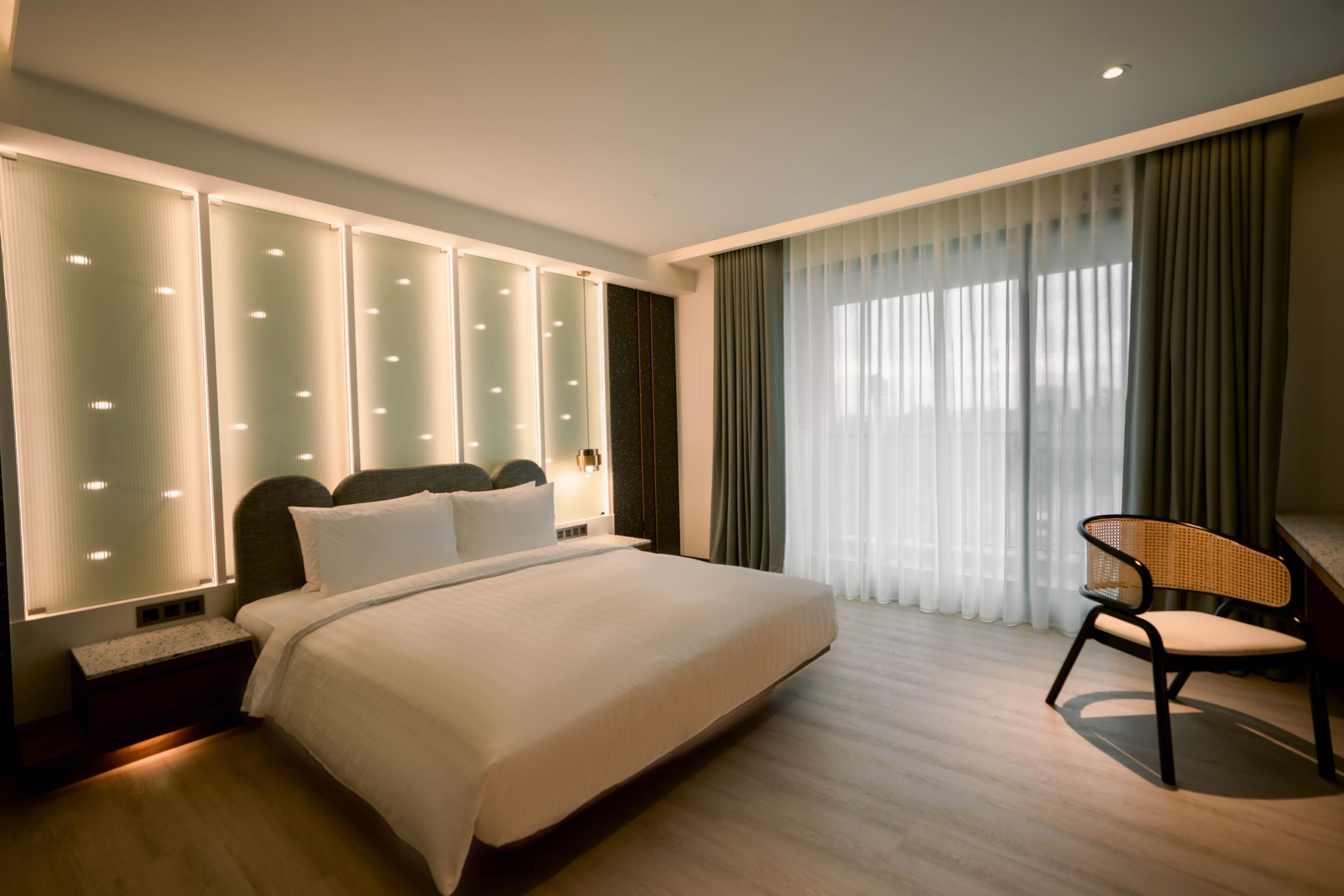 A modern hotel room with a large bed, a nightstand, a chair, and a large window with curtains.