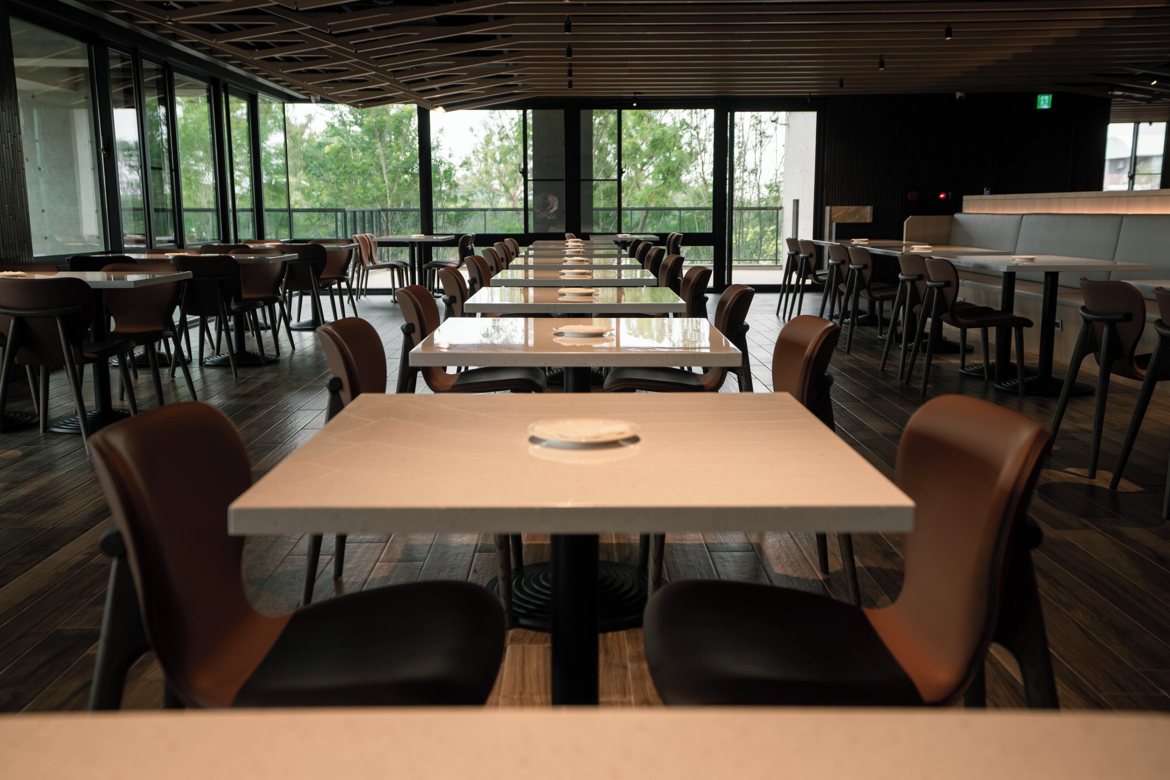 A spacious dining area with long tables and chairs, large windows offering a view of greenery, and a modern ceiling design.