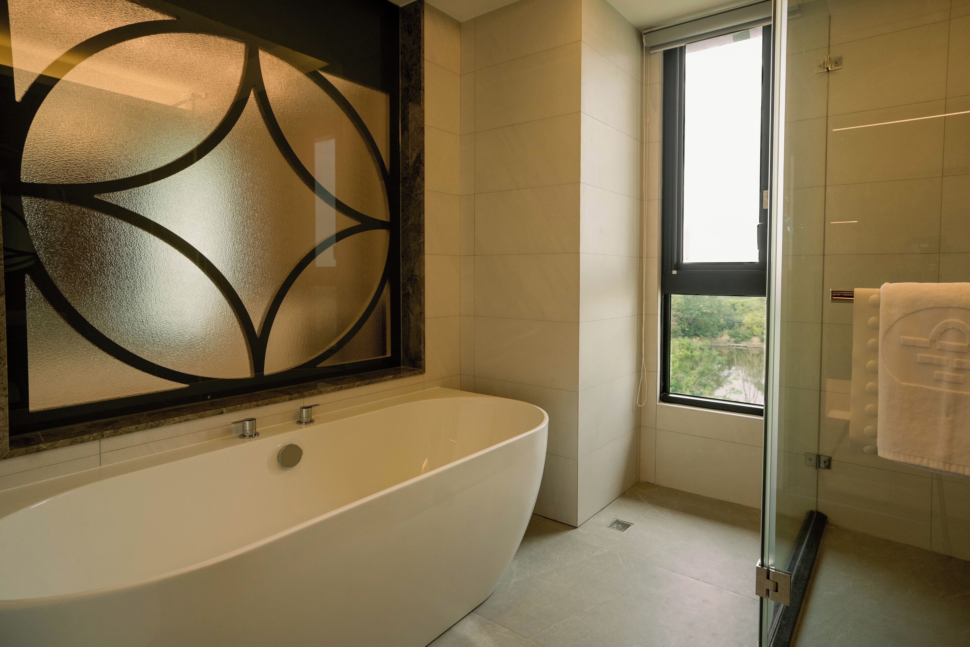 A modern bathroom with a freestanding bathtub, a glass shower enclosure, and a view of greenery through a large window.