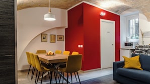 A dining area with a wooden table and yellow chairs, a red accent wall, a blue sofa with a yellow pillow, and a kitchen area with a microwave. - Cozy guesthouse in the center of Rome (Roma)