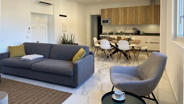 Ein modernes Wohnzimmer mit einem grauen Sofa, einem grauen Sessel und einem Essbereich mit weißen Stühlen und einem Holztisch.