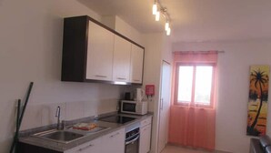 A compact kitchen with white cabinets, a microwave, and a sink. There is a window with a pink curtain and a painting on the wall. - Lovely Pool View 1 bed Apartment Block 6 (Sal)