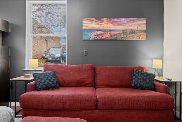 Una sala de estar moderna con un sofá rojo, dos almohadones estampados, un cuadro de paisaje grande y una ventana con persianas.