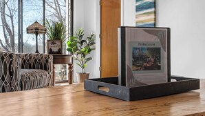 Ein Holztisch mit einem gerahmten Bild von Timbercrest, einer Topfpflanze, einer Lampe und einem gemusterten Sofa in einem Raum mit großen Fenstern und einer Holztür.