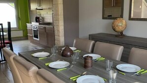 Zona pranzo con un tavolo lungo apparecchiato per quattro, circondato da sedie imbottite. Si intravede in secondo piano una cucina con elettrodomestici moderni.