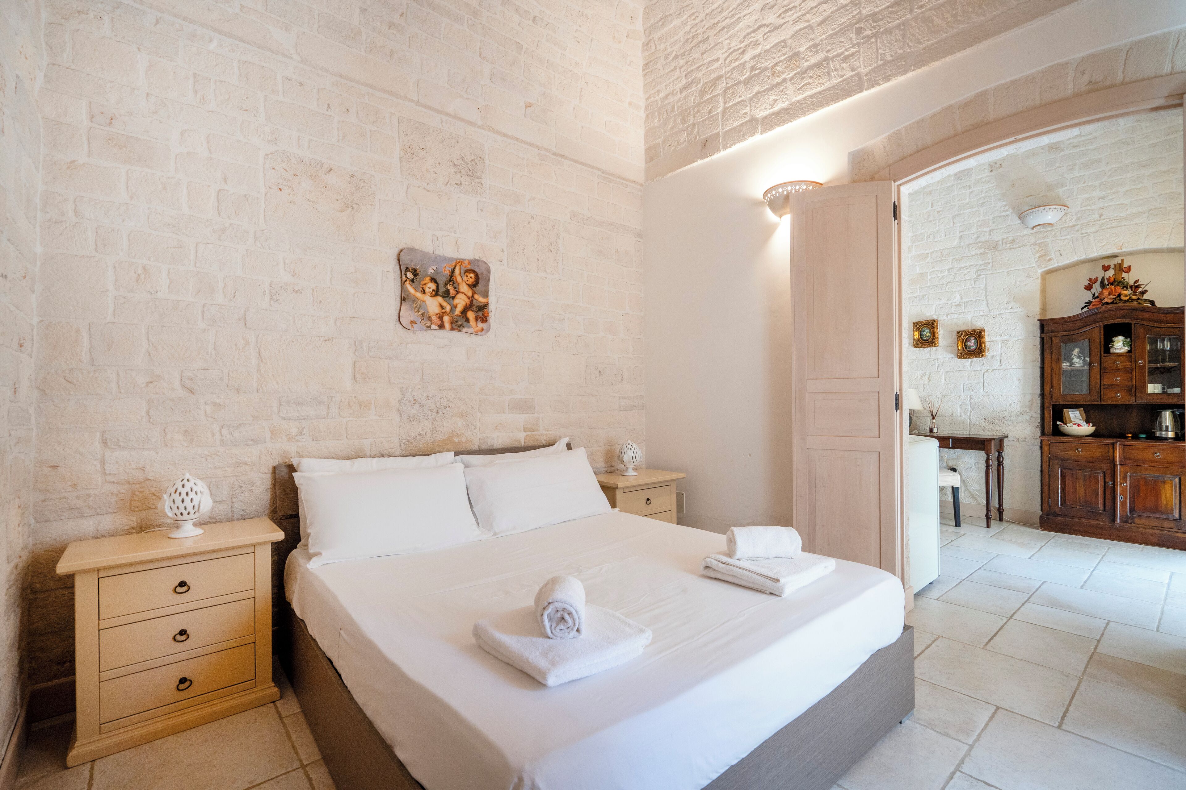 Una cama bien tendida con sábanas blancas, una mesita de noche de madera con cajones, un cuadro colgado en la pared y un mueble de madera con puertas de vidrio.