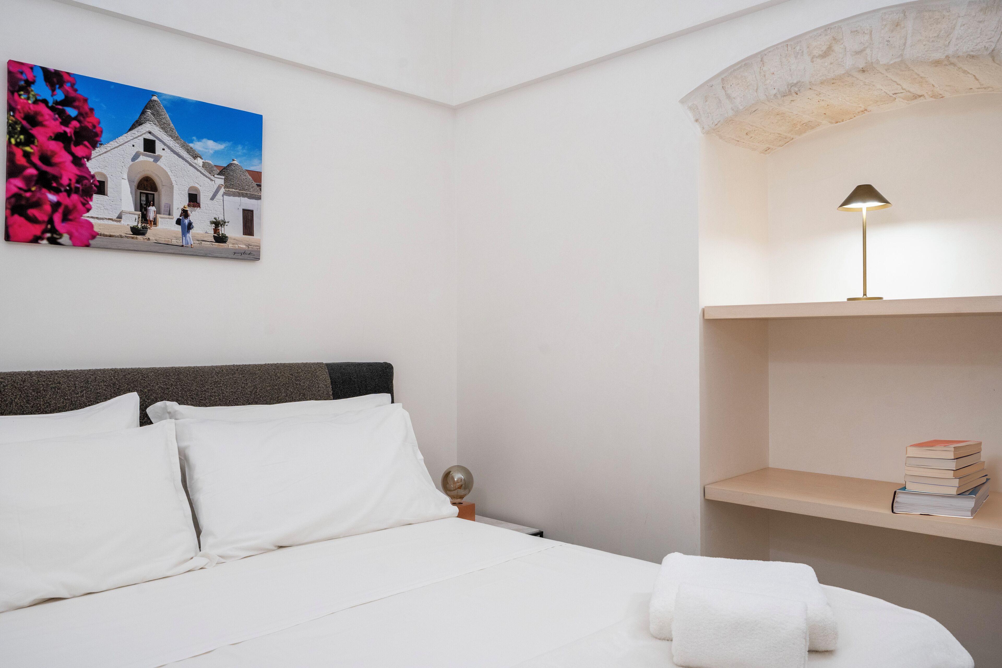 A bedroom with a bed, white pillows, a nightstand with a lamp, a shelf with books, and a painting of a building.