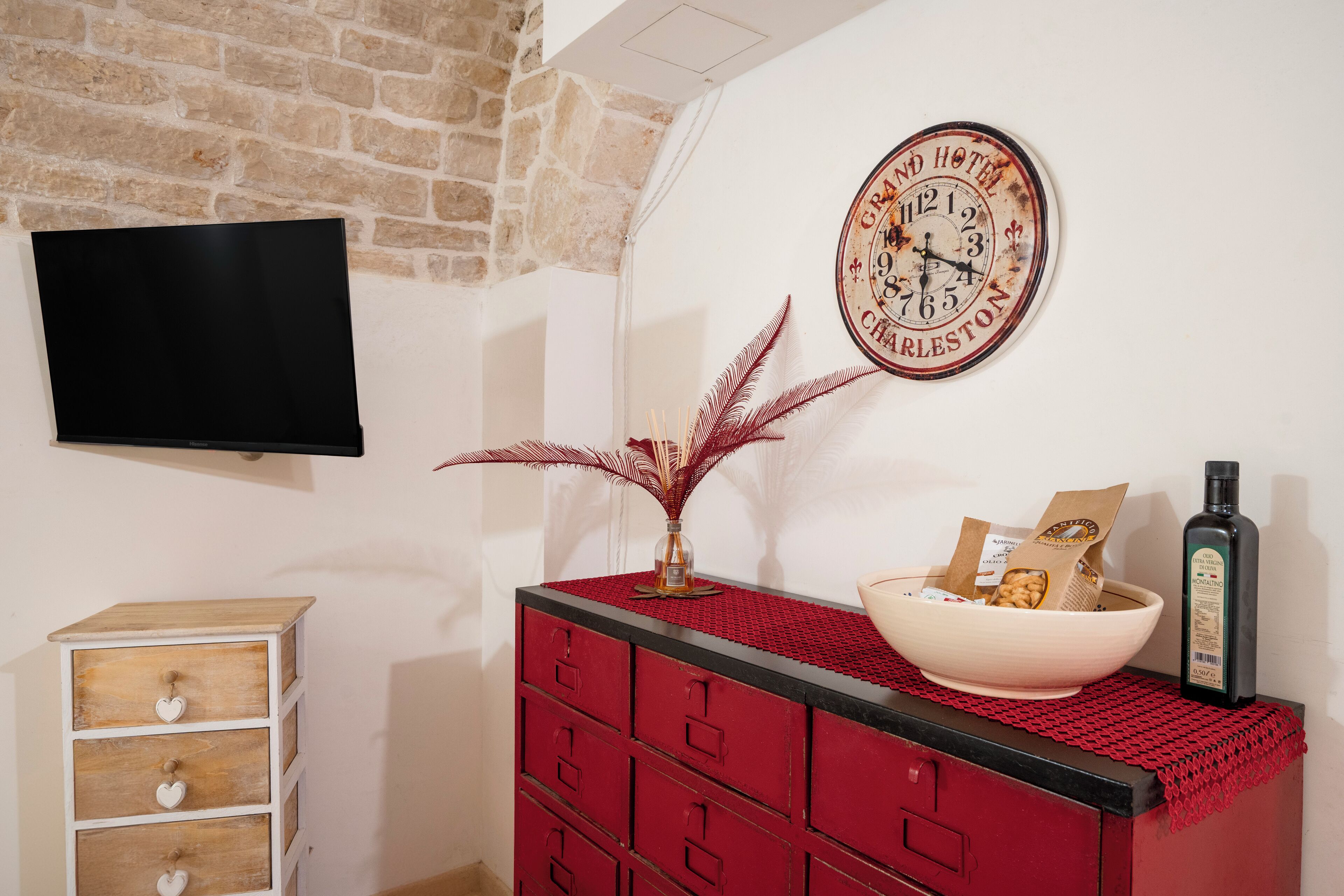 A room with a stone wall, a flat-screen TV, a red dresser with drawers, a bowl on the dresser, a bottle, and a wall clock.