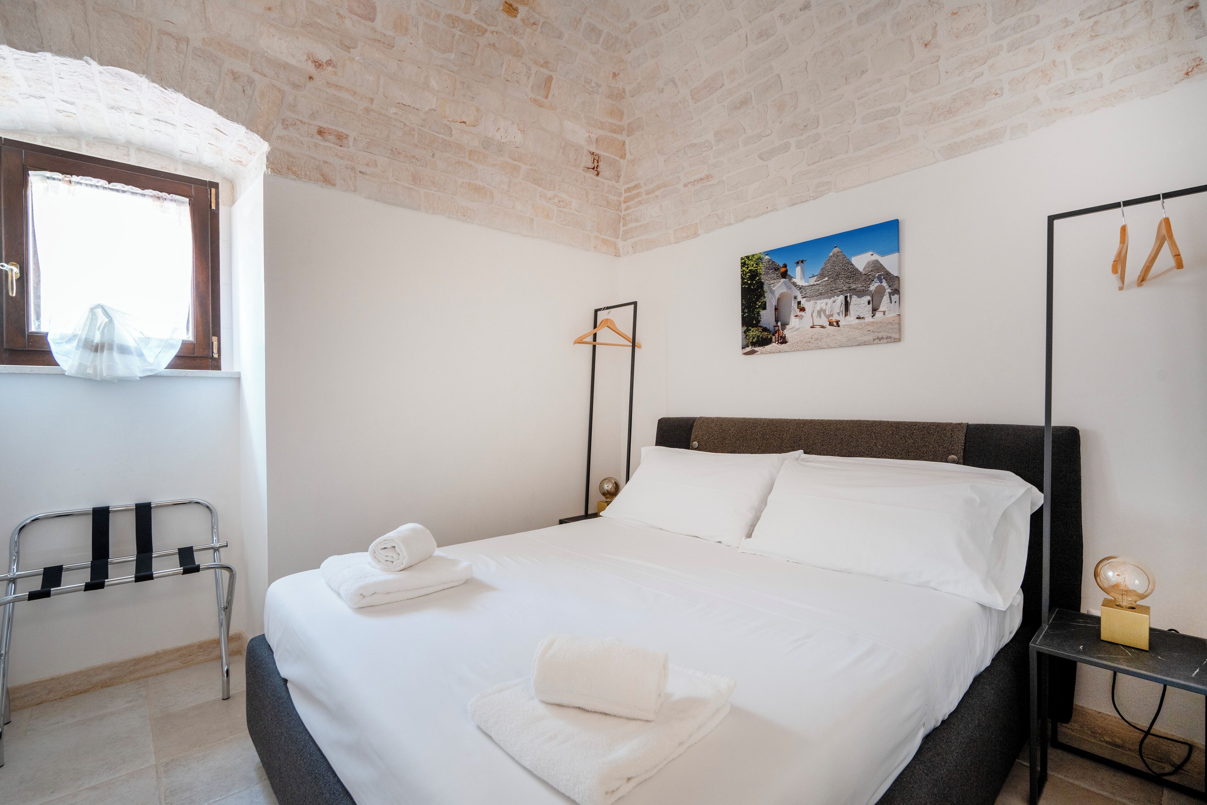 A neatly made bed with white linens and towels, a picture on the wall, and a window with a curtain.