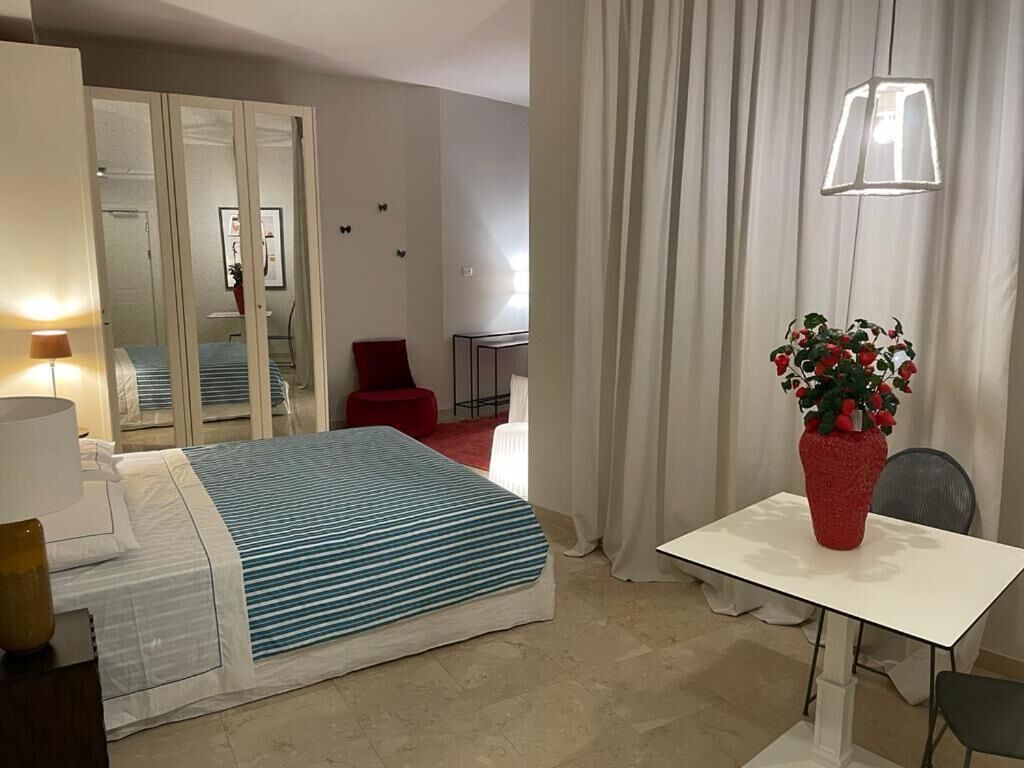 Une chambre d’hôtel avec un lit, un bureau, une chaise, un vase rouge avec des fleurs, une lampe et un miroir.