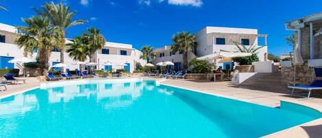 A swimming pool with blue lounge chairs and palm trees, surrounded by white buildings.