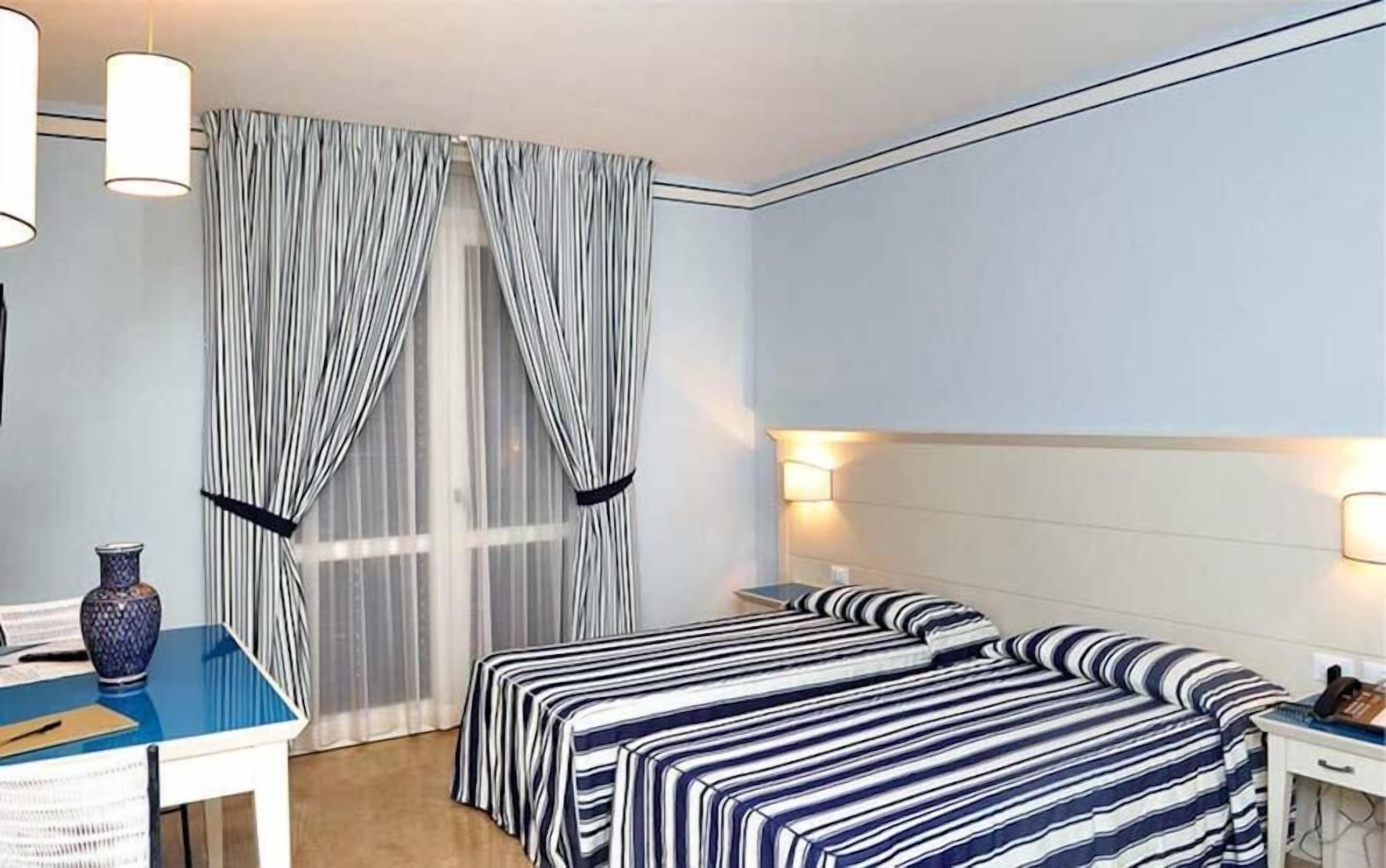 A hotel room with a bed, a desk, and a chair. There are curtains on the window, and a vase on the desk.