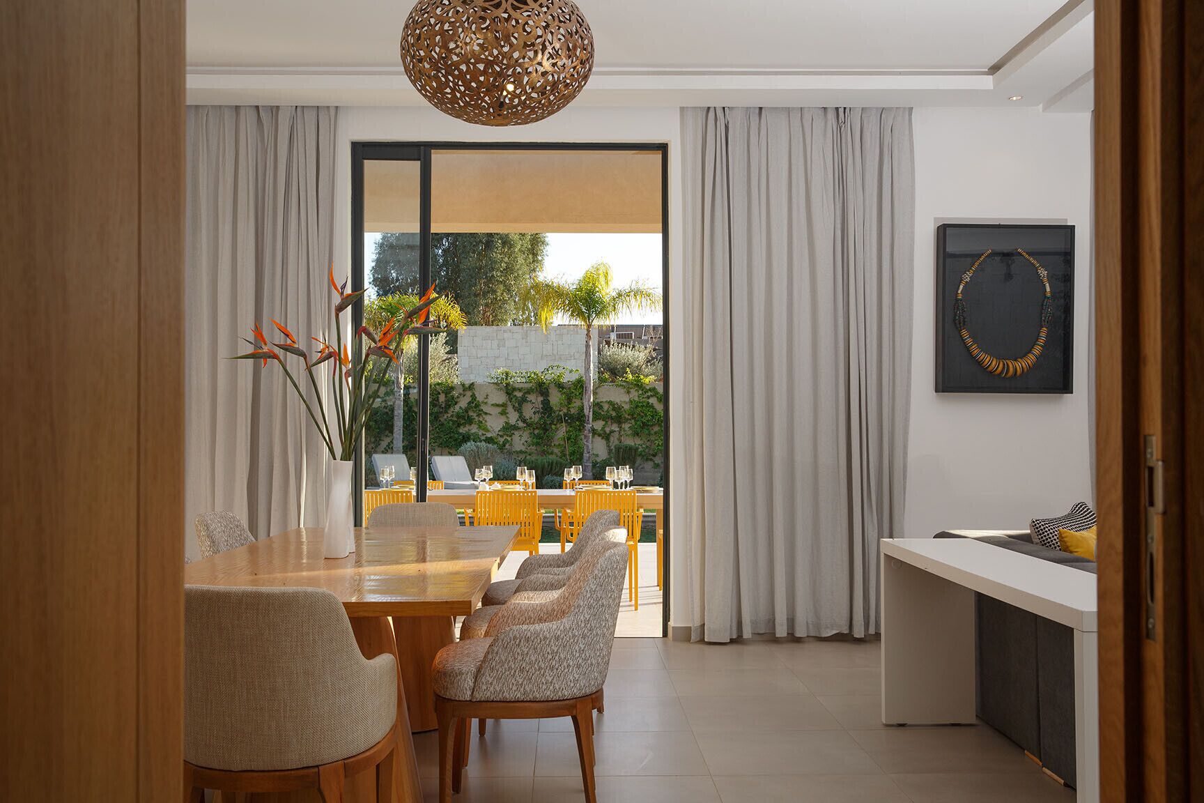 A modern dining area with a wooden table and chairs, a decorative hanging light, and a view of the outdoors.