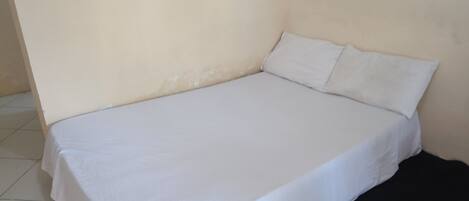 Una cama individual con colchón y almohadas blancas en una habitación con paredes lisas y suelo de baldosas.