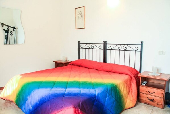 Een bed met een deken in regboogkleuren, een houten nachtkastje met een lamp, en een klein fotolijstje aan de muur.