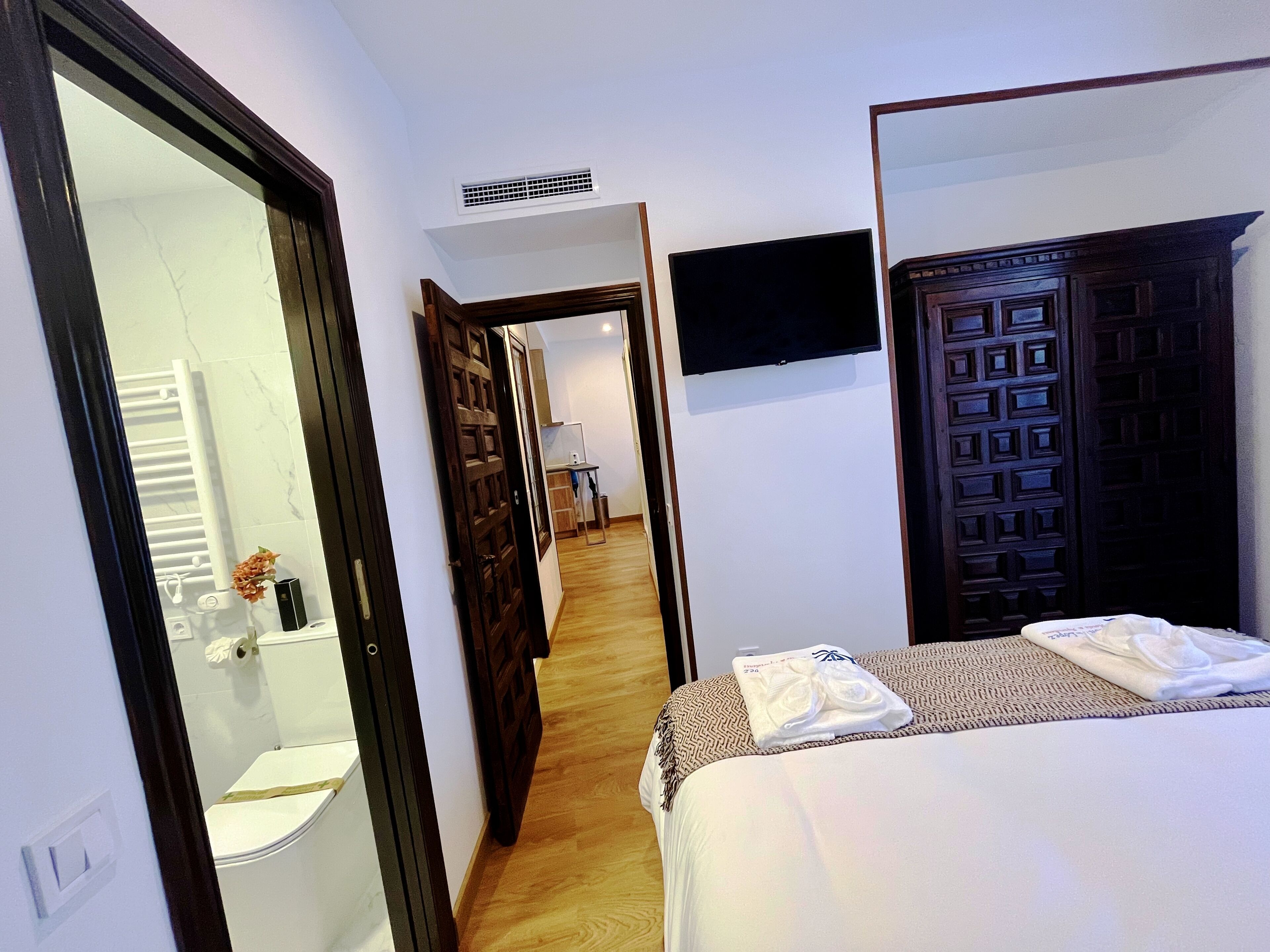 A hotel room with a bed, a TV, a bathroom, and a hallway leading to a kitchen.