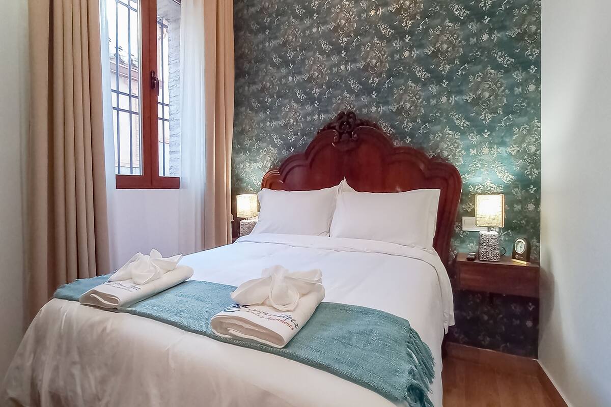 Un lit bien fait, avec des draps blancs et un plaid bleu, deux serviettes blanches pliées sur le lit, une tête de lit en bois et une table de chevet avec une lampe.