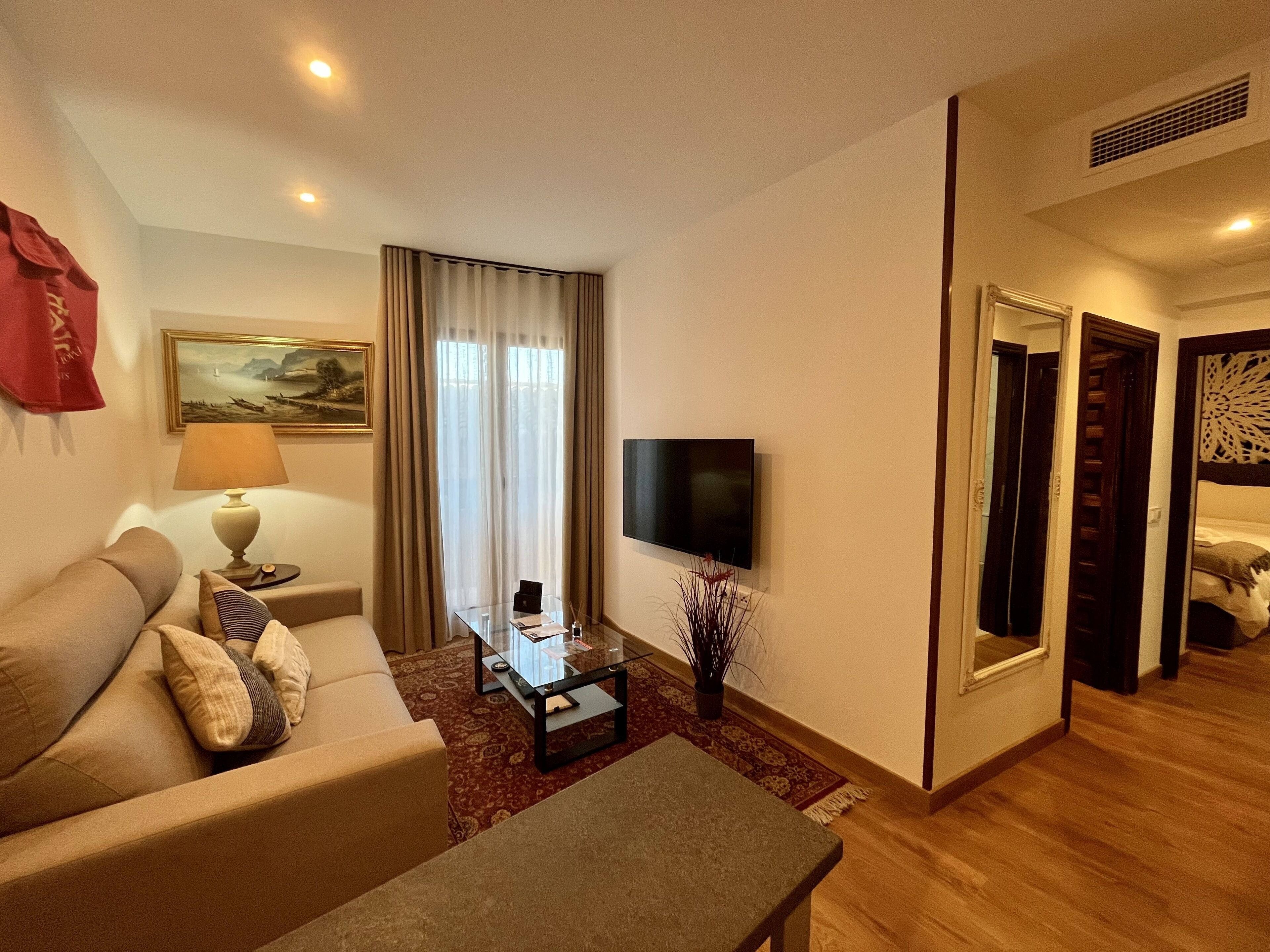 A hotel room with a sofa, a coffee table, a TV, a painting, and a window with curtains.